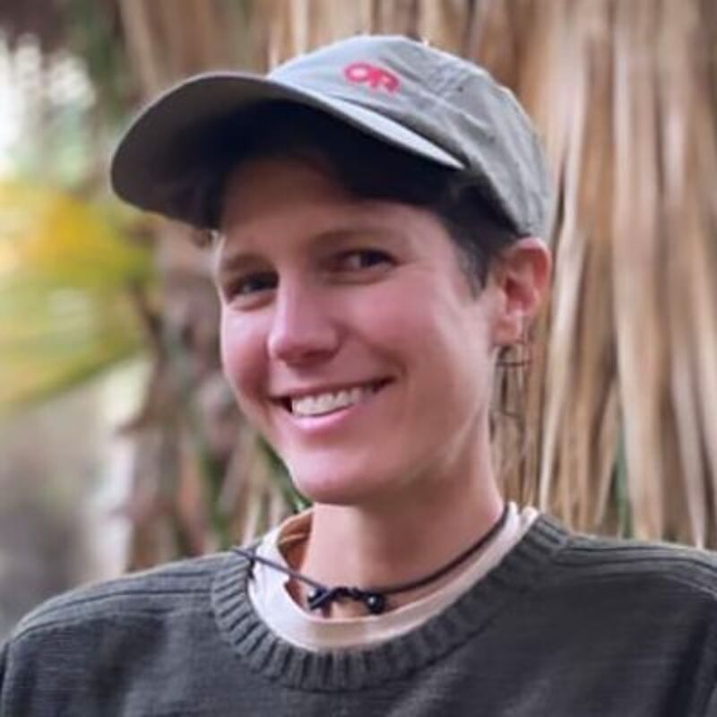 The image shows a person with short hair wearing a cap and a sweater. The person is smiling and looking at the camera. The background is blurred, but it seems to be outdoors, possibly in a park or natural setting. The overall impression is of a casual and friendly portrait.